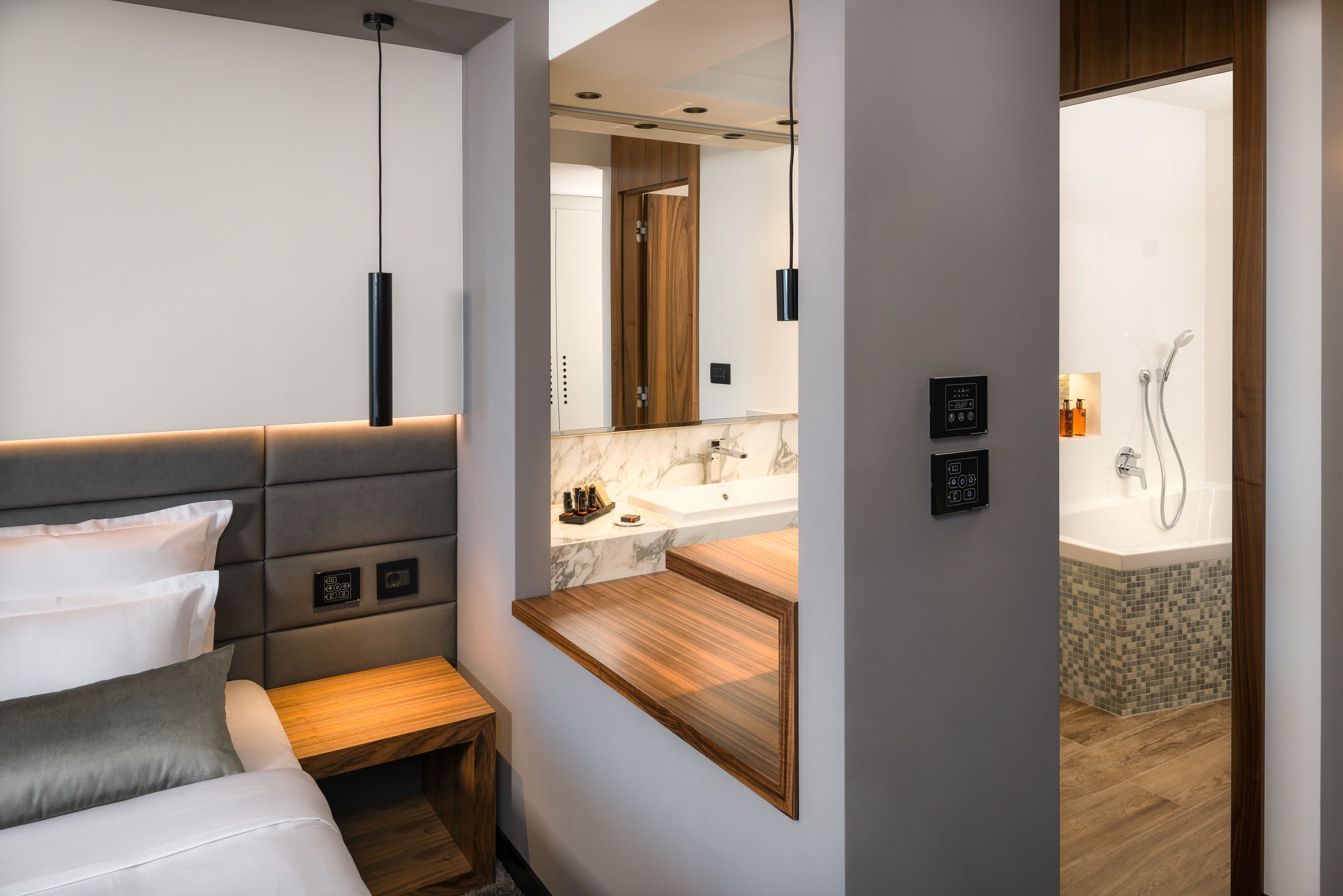 Una habitación de hotel moderna con cama, mesita de noche, banco empotrado y baño con ducha, lavabo y espejo.