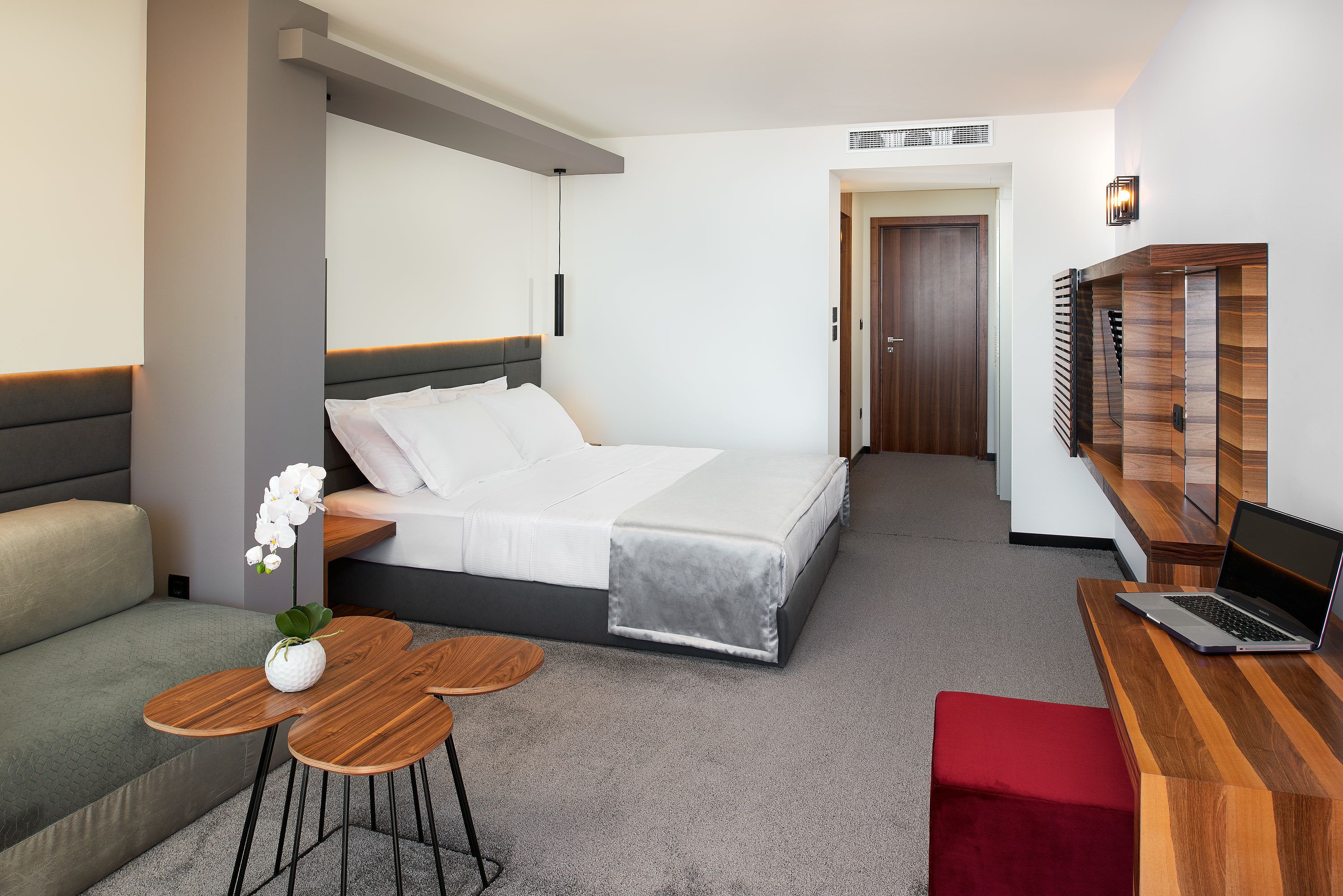 A modern hotel room with a bed, a desk with a laptop, a small table with a vase, and a red bench.