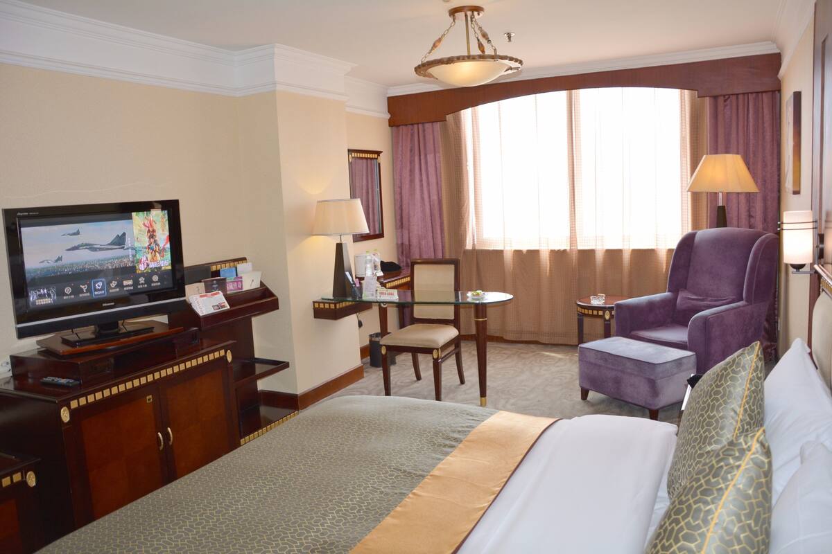 Une chambre d’hôtel comprenant un lit, une télévision, un bureau avec une chaise, un fauteuil violet et une fenêtre avec des rideaux.