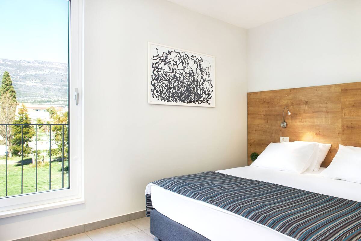 A bedroom with a large bed, a wooden headboard, a view of a balcony, and a piece of abstract art on the wall.