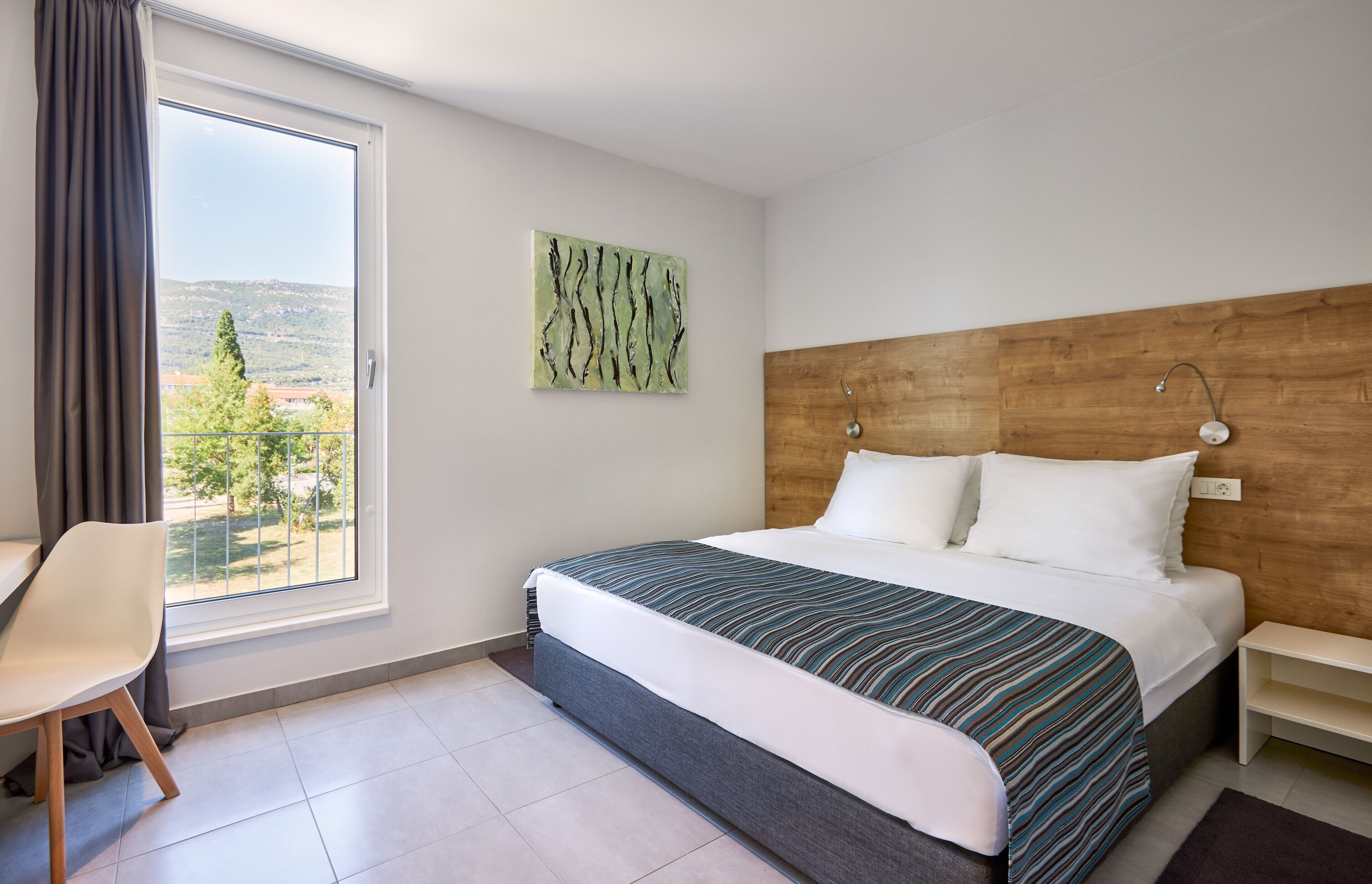 A hotel room with a large bed, a chair, a wooden headboard, a painting, and a view of the outdoors.