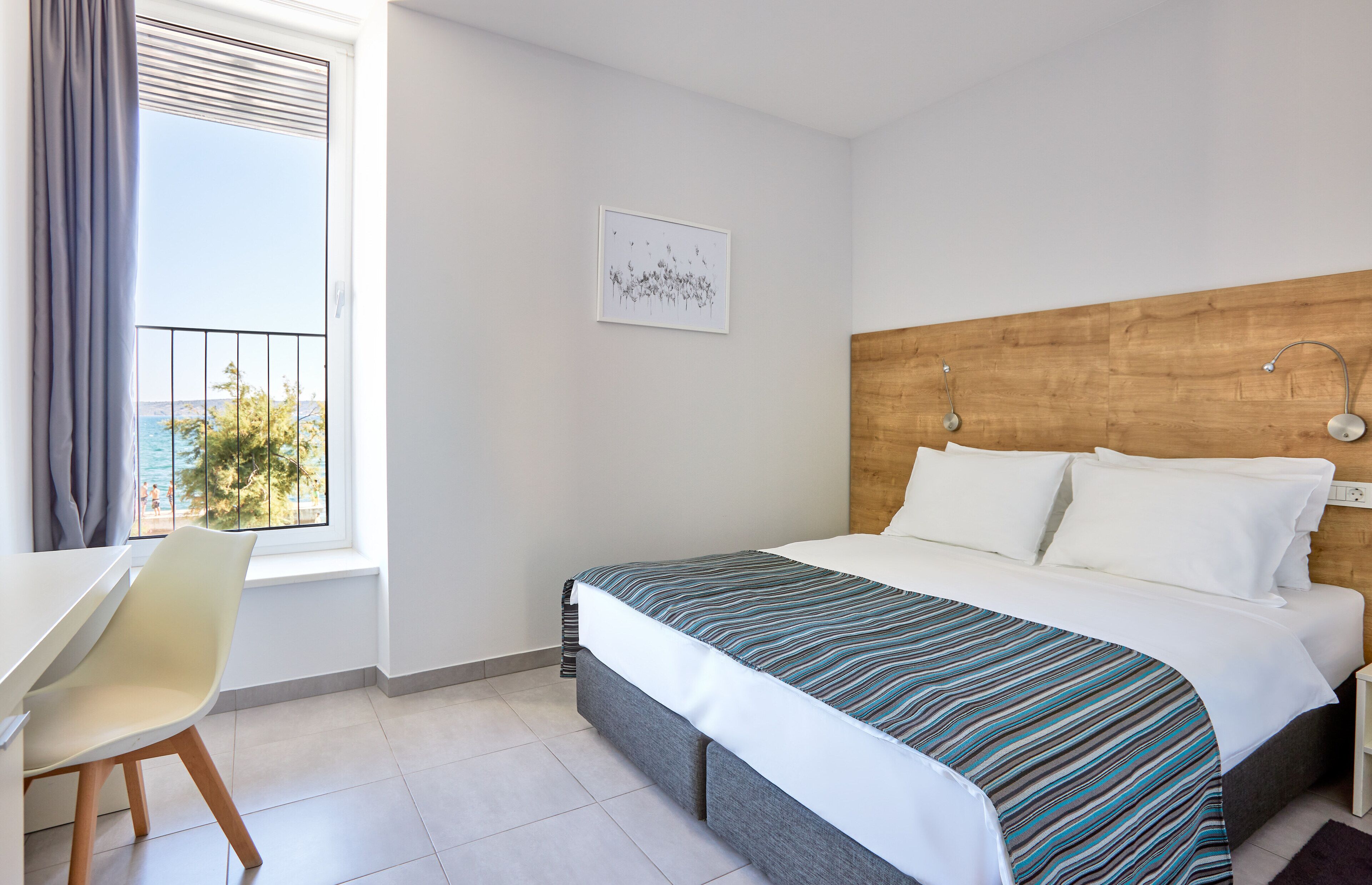 A hotel room with a bed, a desk, a chair, a window with a view of the sea, and a framed picture on the wall.