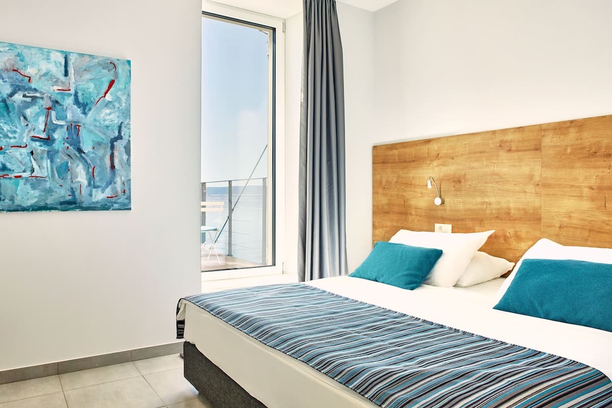 A modern hotel room with a large window, a wooden headboard, and a blue abstract painting on the wall.