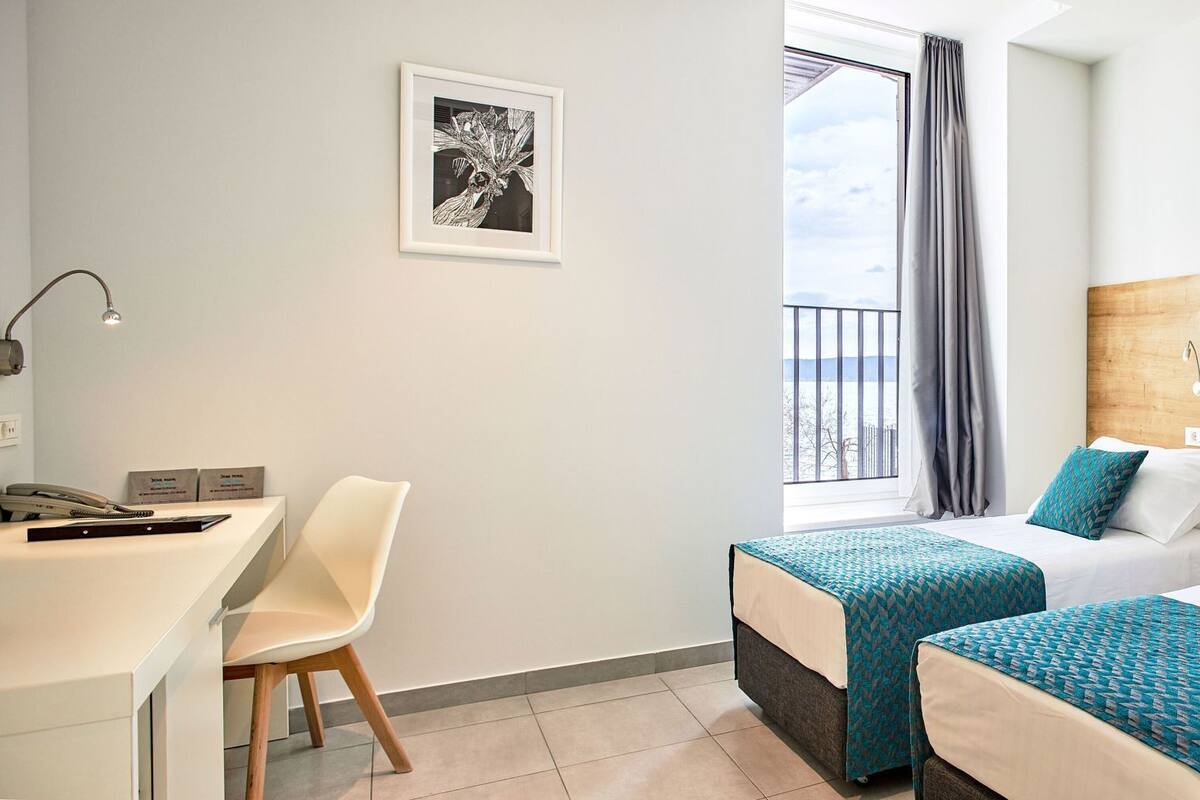 A hotel room with two beds, a desk, a chair, a framed picture on the wall, and a window with curtains.