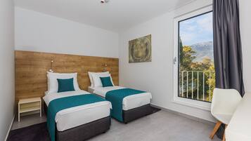 Habitación de hotel con dos camas, cabecera de madera, un ventanal con vistas, una silla blanca y una mesita.
