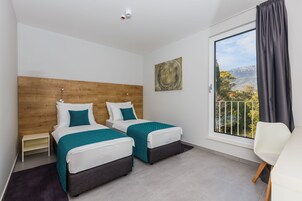 Habitación de hotel con dos camas, cabecera de madera, un ventanal con vistas, una silla blanca y una mesita.
