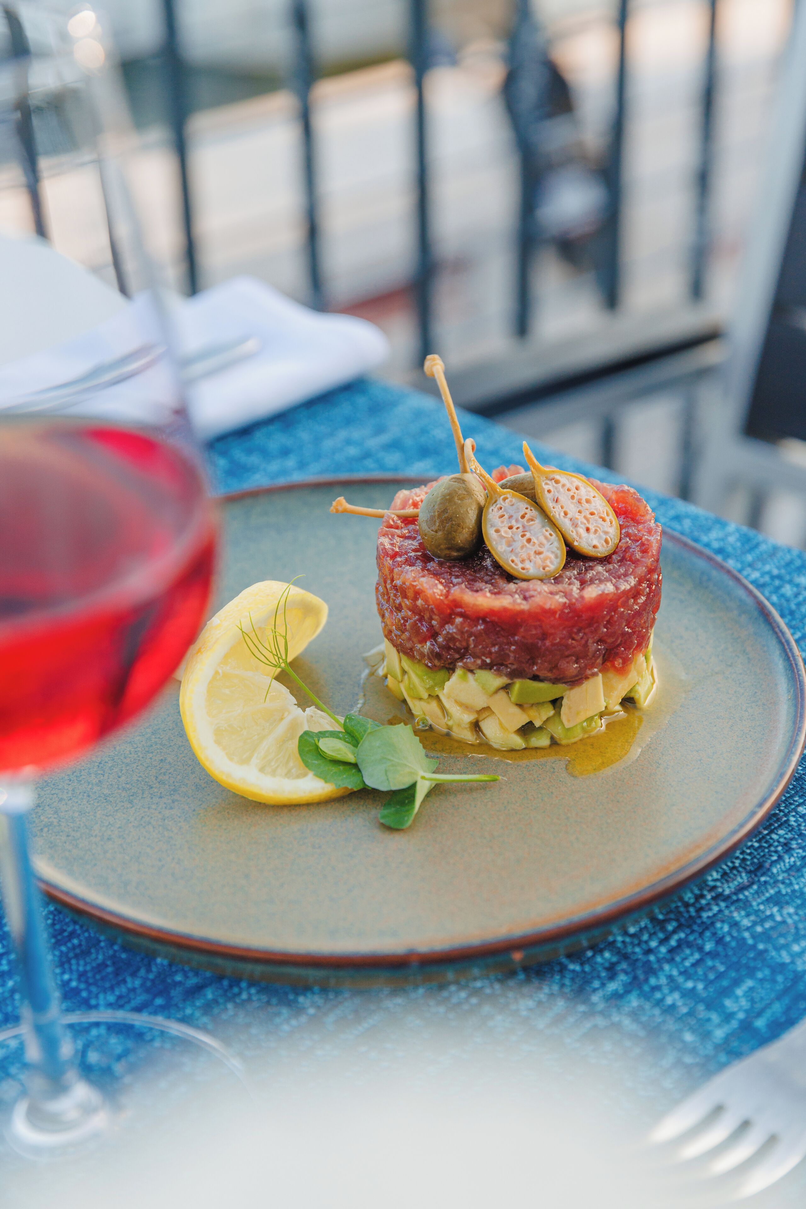 A plate of steak tartare with lemon and garnish, a glass of red wine, and a blue tablecloth.