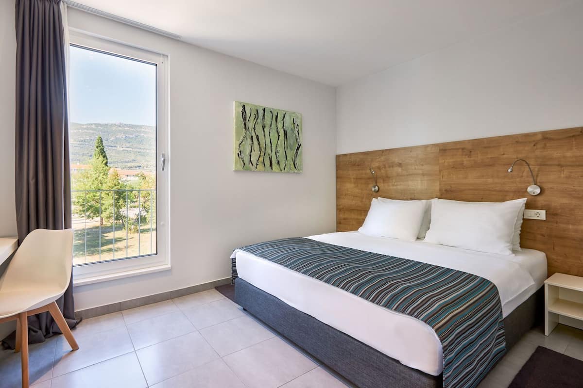 A hotel room with a bed, a chair, a nightstand, a painting, and a view of the outdoors.
