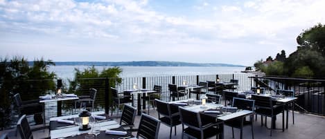 An outdoor dining area with tables and chairs, overlooking a body of water and a distant shoreline.