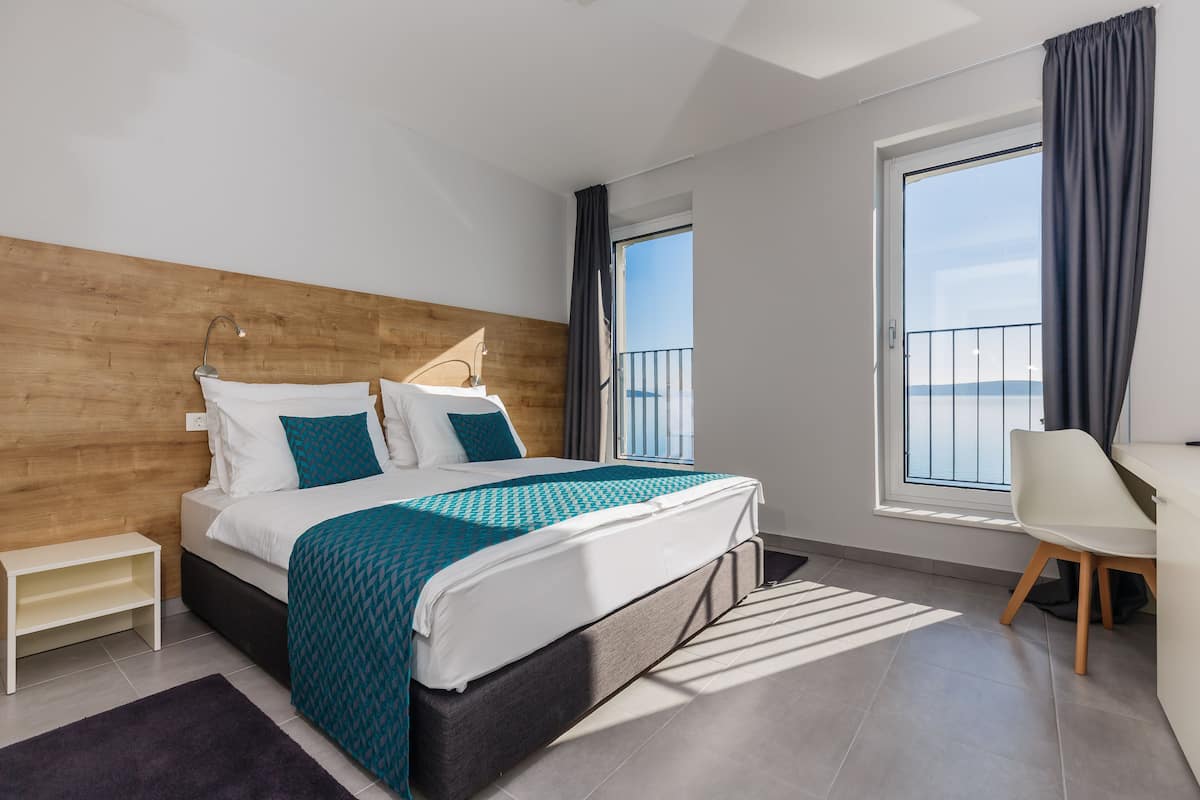 A modern hotel room with a large bed, a desk, a chair, a small table, and a balcony with a view.