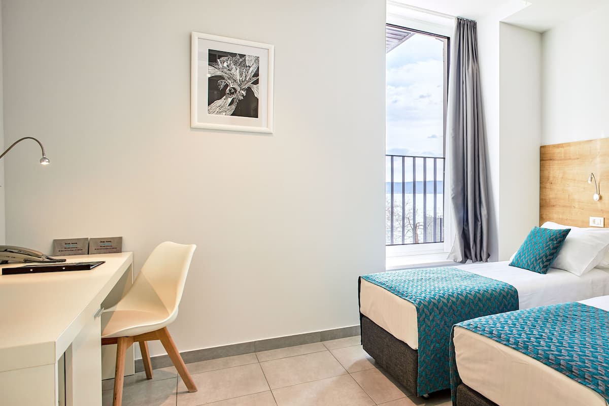 A hotel room with two beds, a desk, a chair, a framed artwork, and a view of the outdoors.