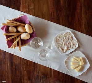 Una mesa de madera cubierta con un mantel blanco, una cesta de palitos de pan, un plato de pescado y un plato con lonchas de queso.