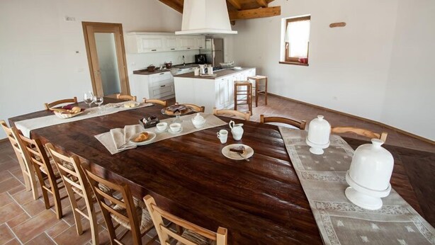 Een ruime eethoek met een grote houten tafel, witte kastjes en een keuken op de achtergrond.