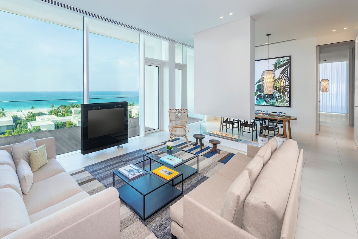 A modern living room with a large TV, comfortable seating, a glass coffee table, and a view of the ocean.