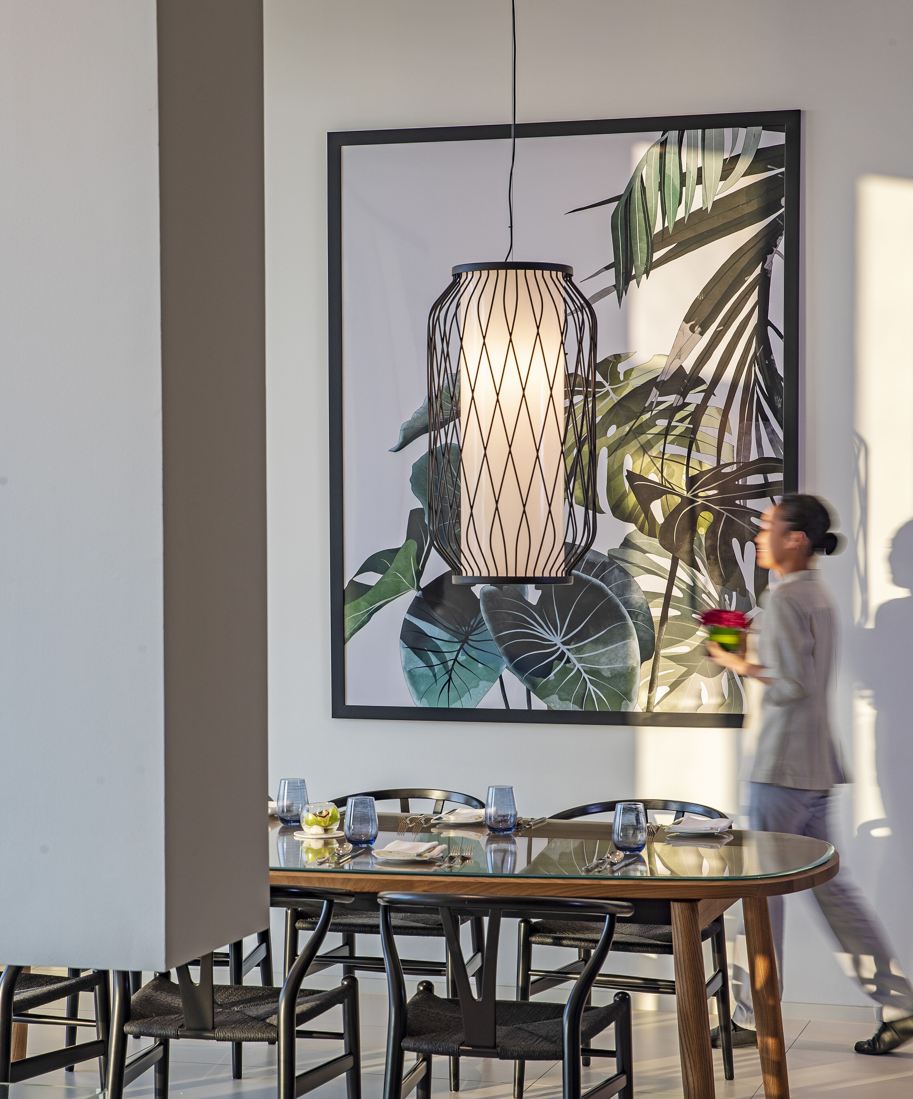 A dining area with a table set for four, surrounded by chairs with metal frames and cushioned seats. A large framed artwork depicting leaves and a hanging light fixture is visible on the wall.