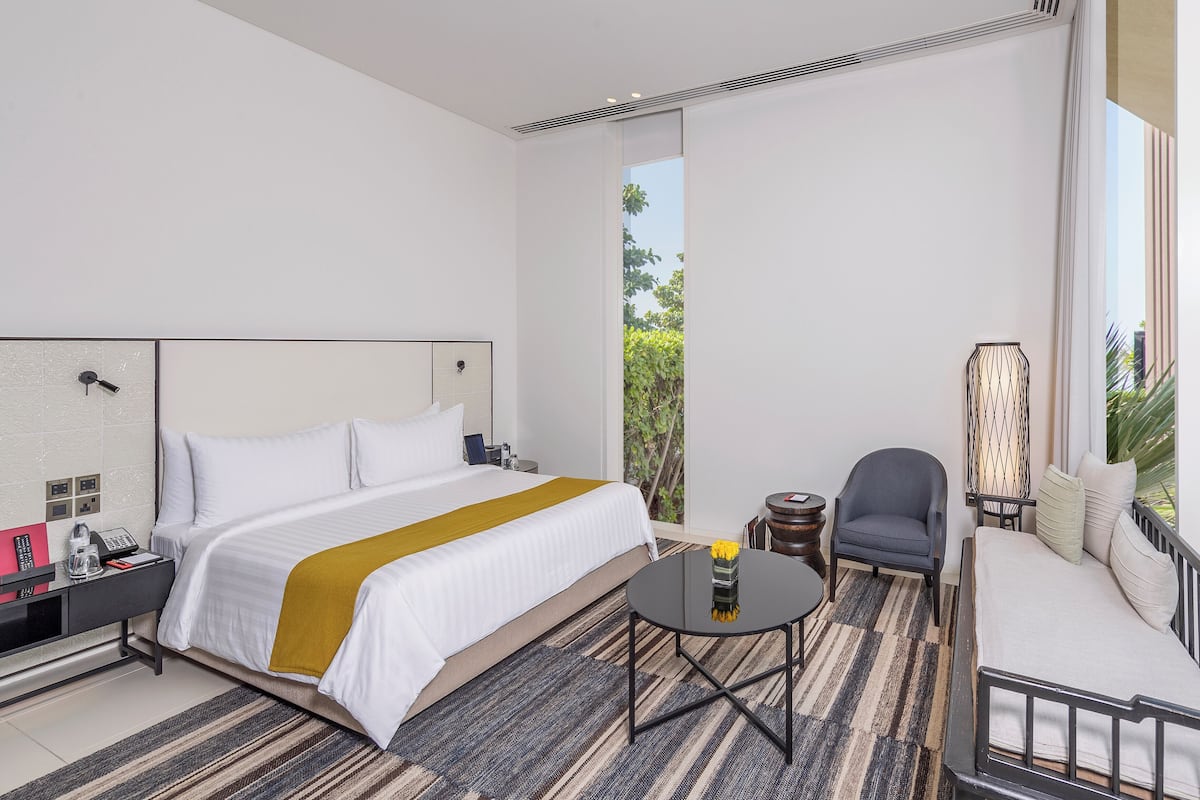 A hotel room with a bed, a small round table with a vase, a chair, a sofa, and a large window with a view of greenery.