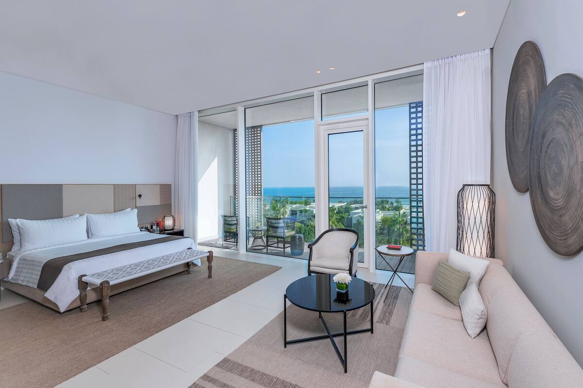 A modern hotel room with a large bed, a sofa, a coffee table, and a balcony with a city view.