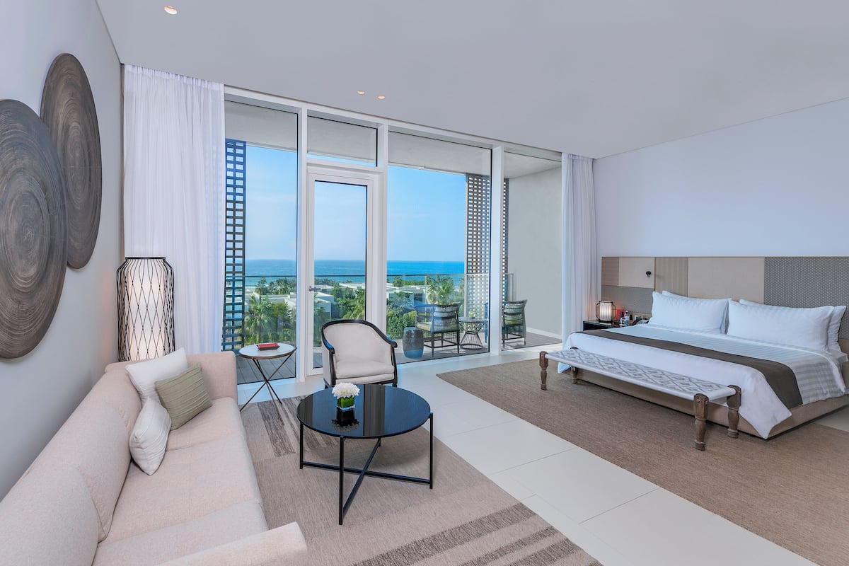 A modern hotel room with a large bed, a sofa, a small table, and a balcony with a view of the ocean.