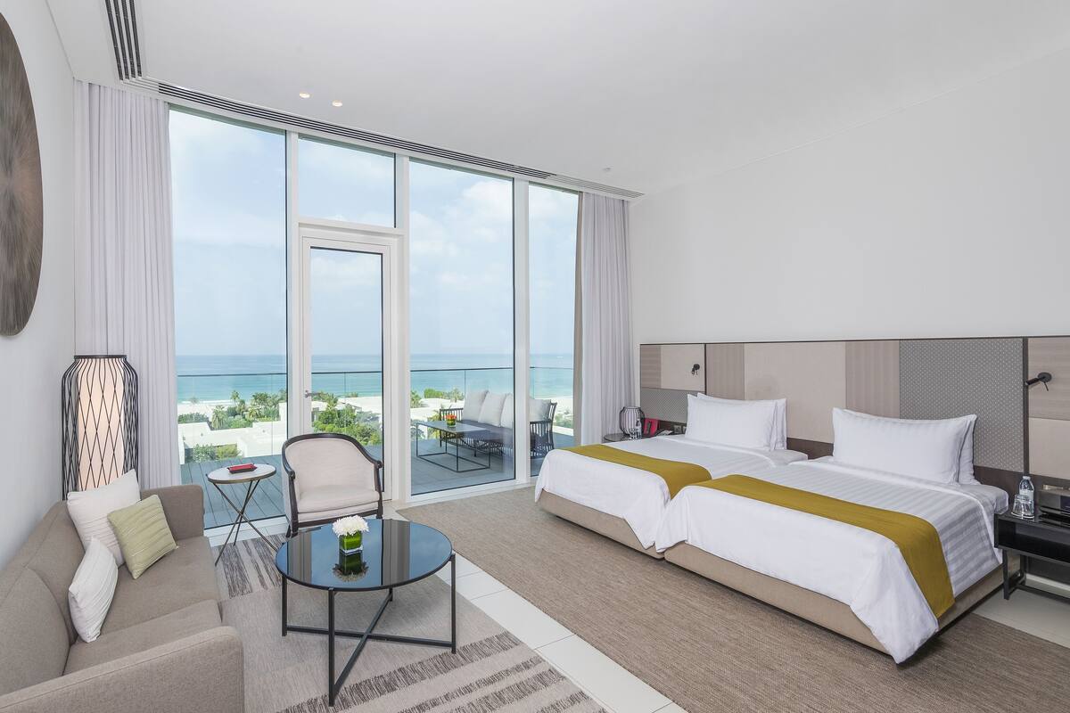 A modern hotel room with a large bed, a sofa, a small table, and a balcony with a view of the ocean.
