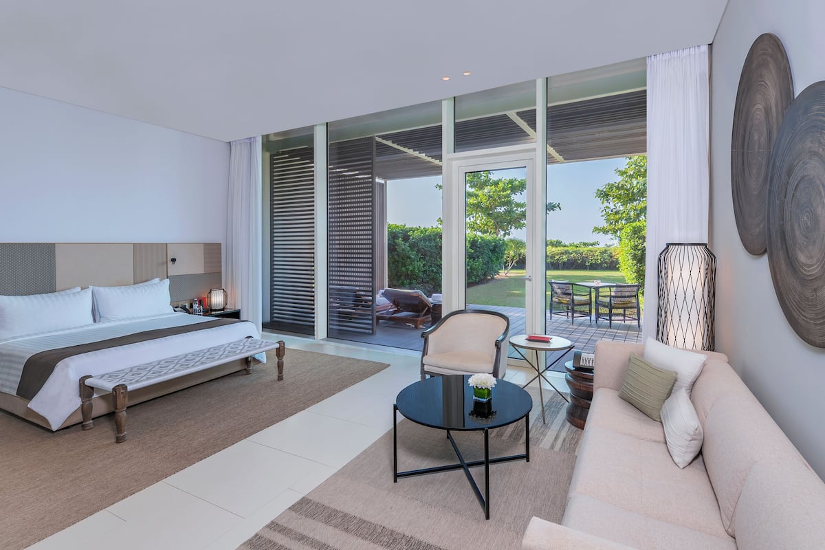 A modern hotel room with a large bed, a sofa, a small table, and a view of the garden through the glass door.