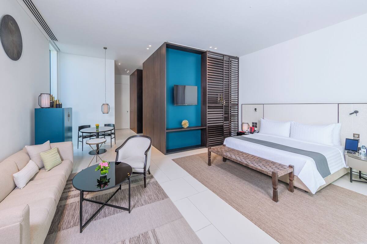 A modern hotel room with a bed, a sofa, a dining table, and a TV.