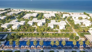 Ett semesterkomplex med flera byggnader, ett poolområde med solstolar och utsikt över stranden.