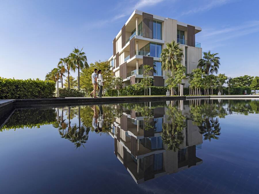 A modern building with a reflection in the water, surrounded by palm trees and greenery.