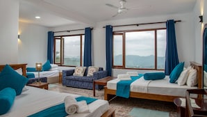Habitación de hotel con dos camas, un sofá, ventilador de techo y ventanas grandes con cortinas.