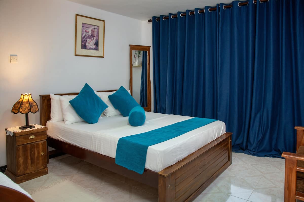A neatly made bed with blue bedding, a wooden nightstand with a lamp, a mirror, and a blue curtain.