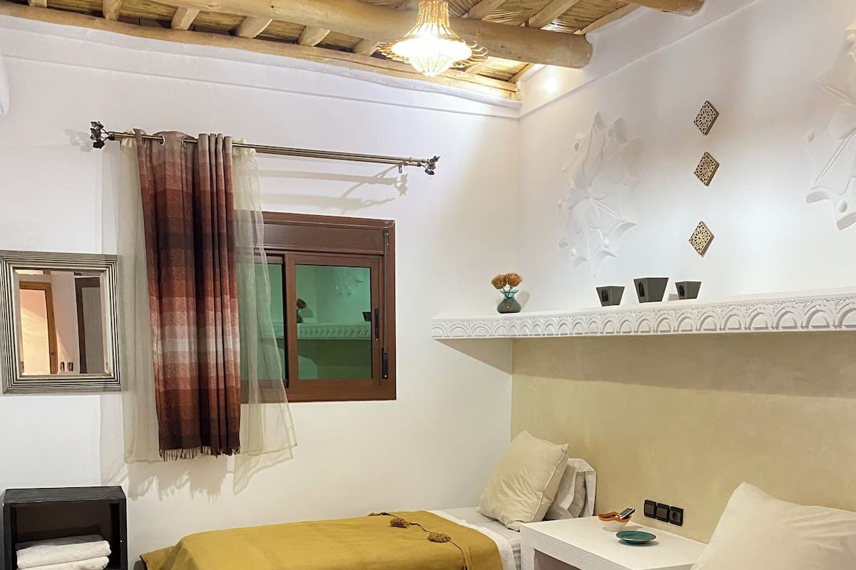 Une chambre avec deux lits, une petite table, une chaise, une étagère avec des objets décoratifs et une fenêtre avec des rideaux.