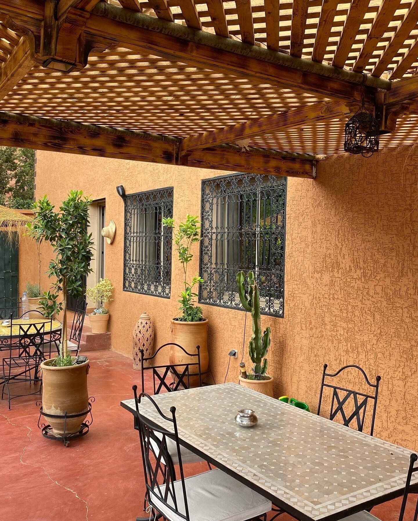 An outdoor patio with a table and chairs, surrounded by potted plants and a wooden pergola.
