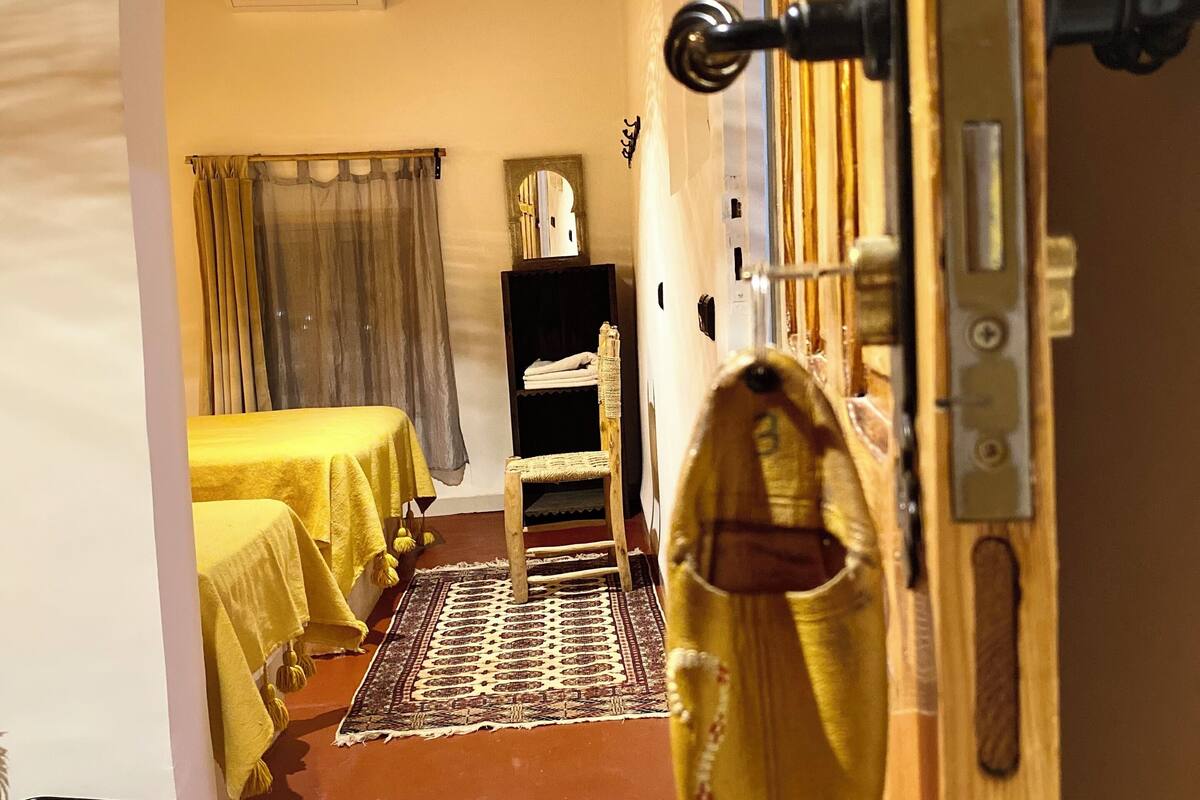 Un couloir étroit avec une porte en bois, une petite chaise en bois et un lit recouvert d’une couverture jaune.