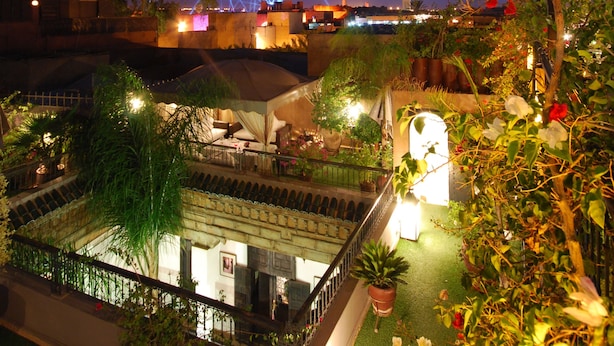 Un jardin sur le toit, agrémenté de plantes en pot, un petit espace aménagé pour s’asseoir et une vue sur les toits de la ville au crépuscule.