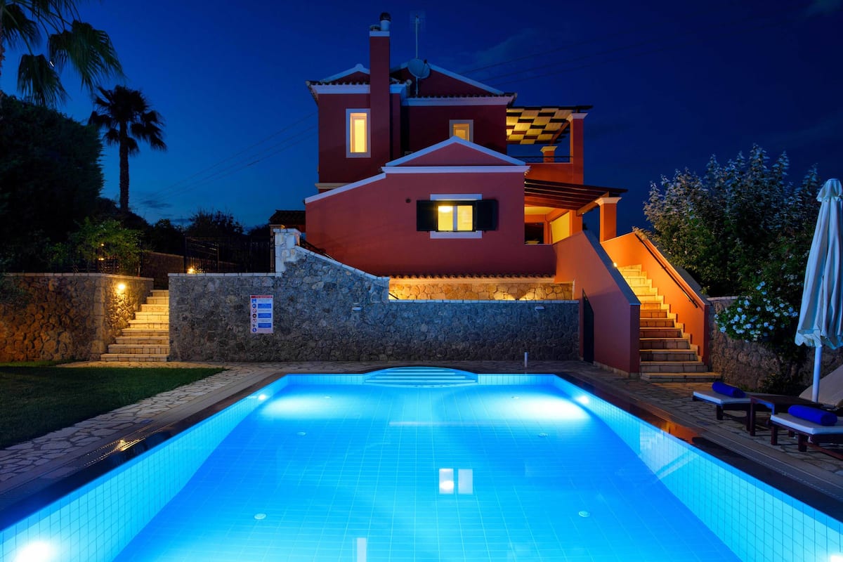 A swimming pool with a stone wall and a red house with a balcony.