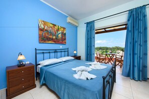 A bedroom with a bed, a nightstand, a lamp, a painting, and a view of the outdoors. - Il Tesori Dassia Villas (Corfu)