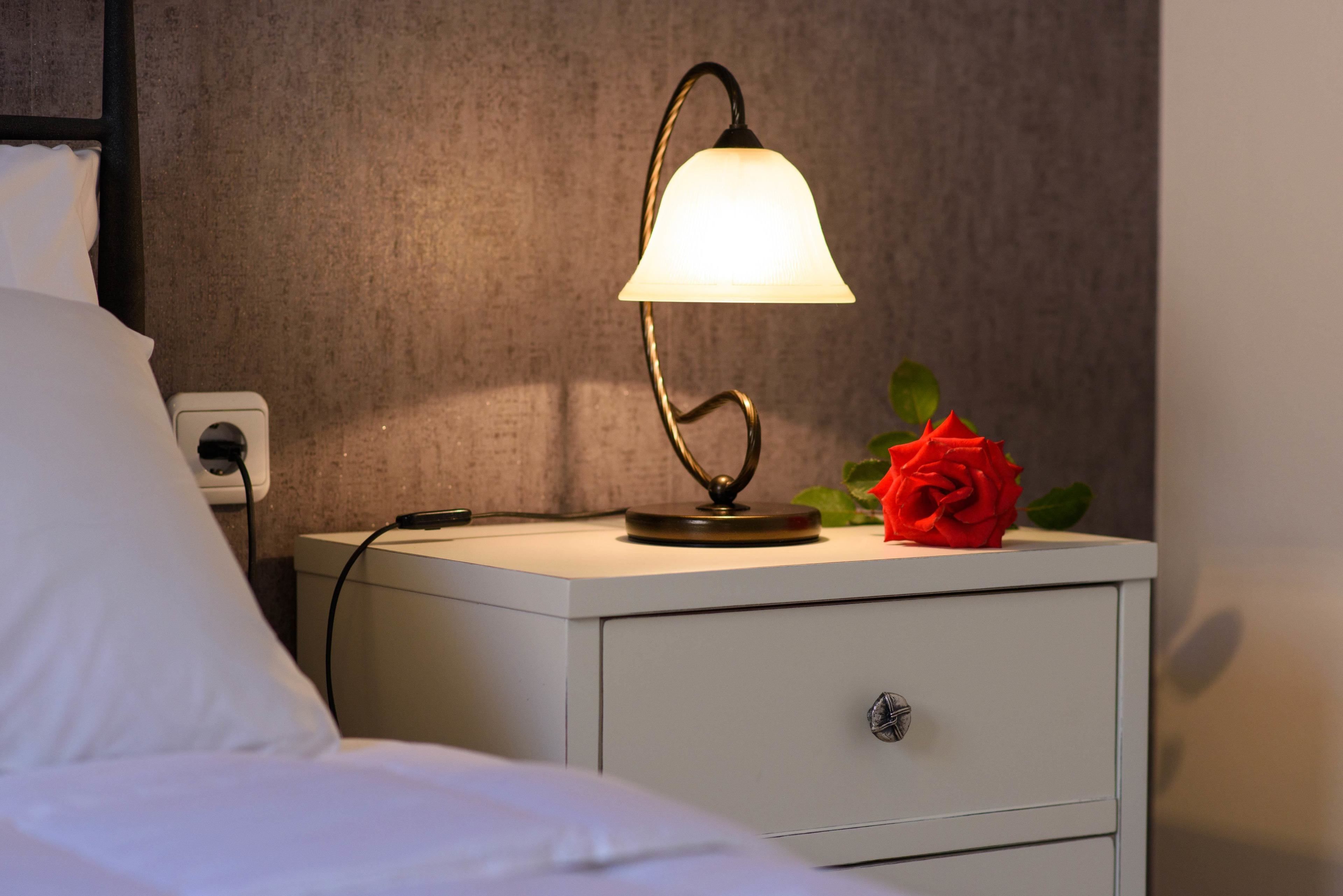 A neatly made bed with white linens, a bedside table with a lamp, a red rose, and a power outlet.