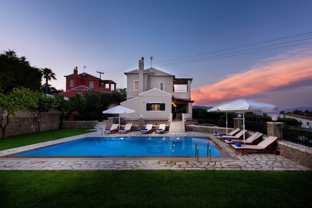 A swimming pool with lounge chairs and umbrellas, surrounded by a stone patio and a house in the background.