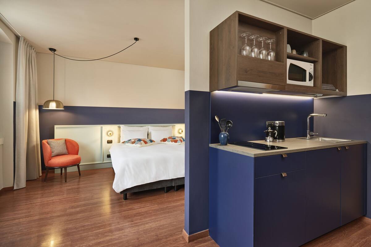 A modern hotel room with a bed, a small orange chair, a kitchenette with a microwave, and a coffee maker.