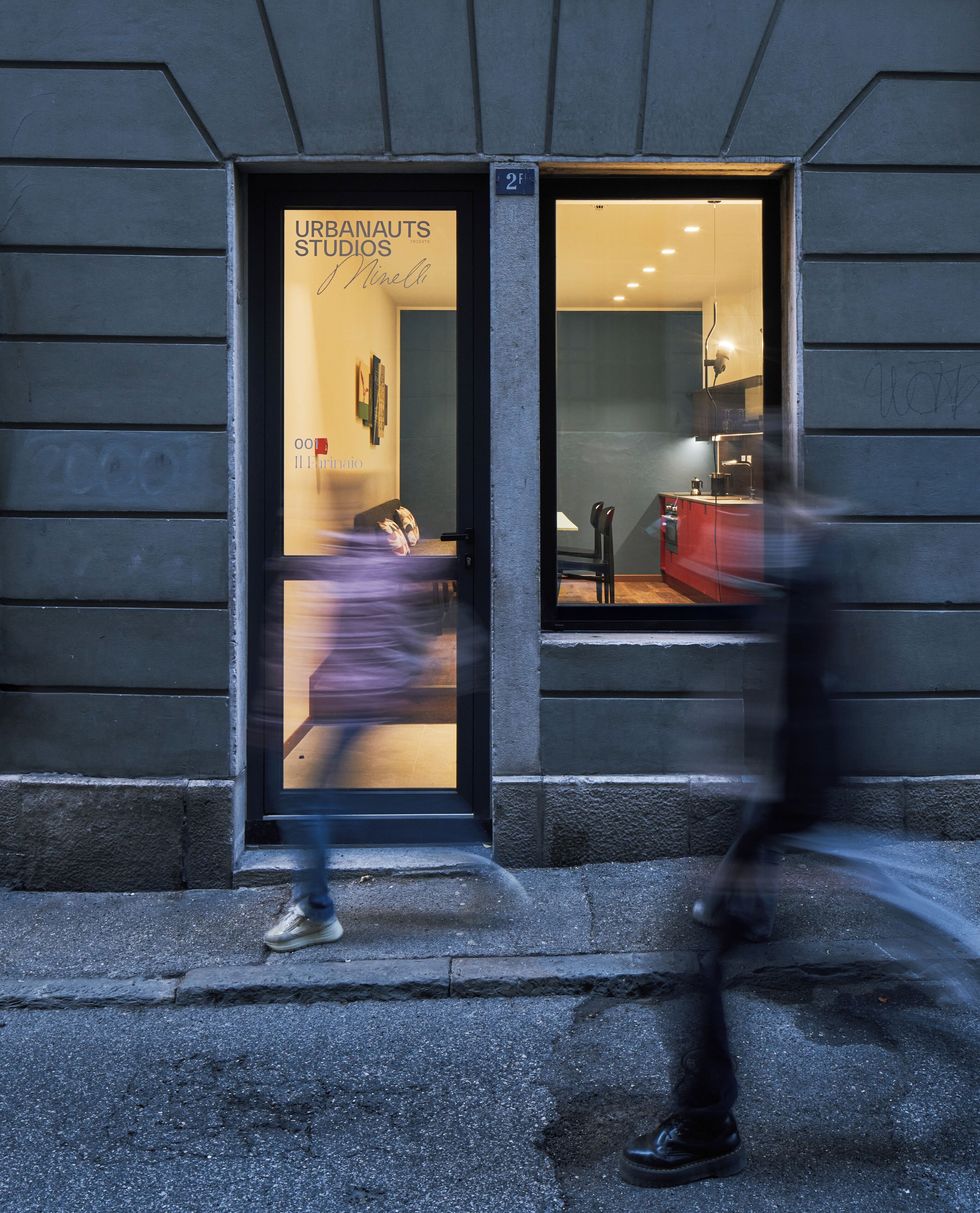 A building entrance with a glass door labeled 'URBANAUTS STUDIOS' and a visible interior with a red counter and seating area.