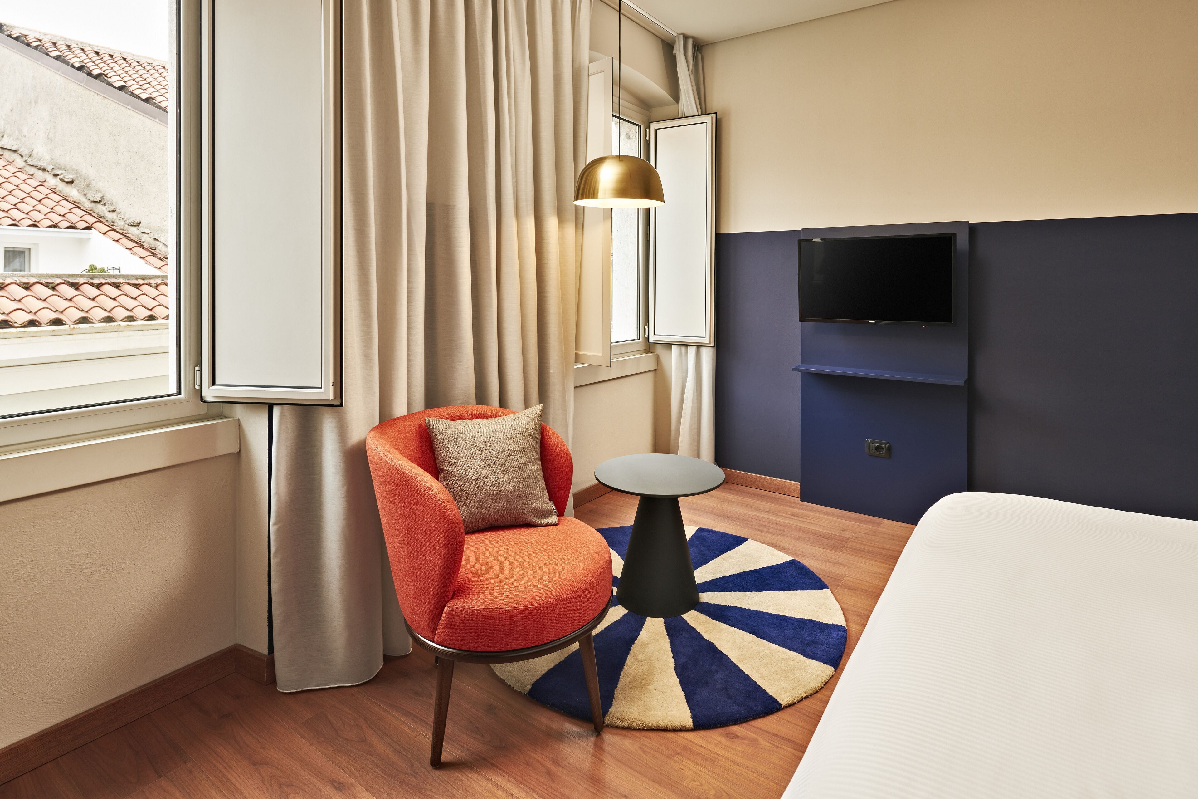 Una camera d'albergo con un letto grande, un tavolino rotondo, una poltrona rossa, una lampada a sospensione dorata e un televisore montato a parete.