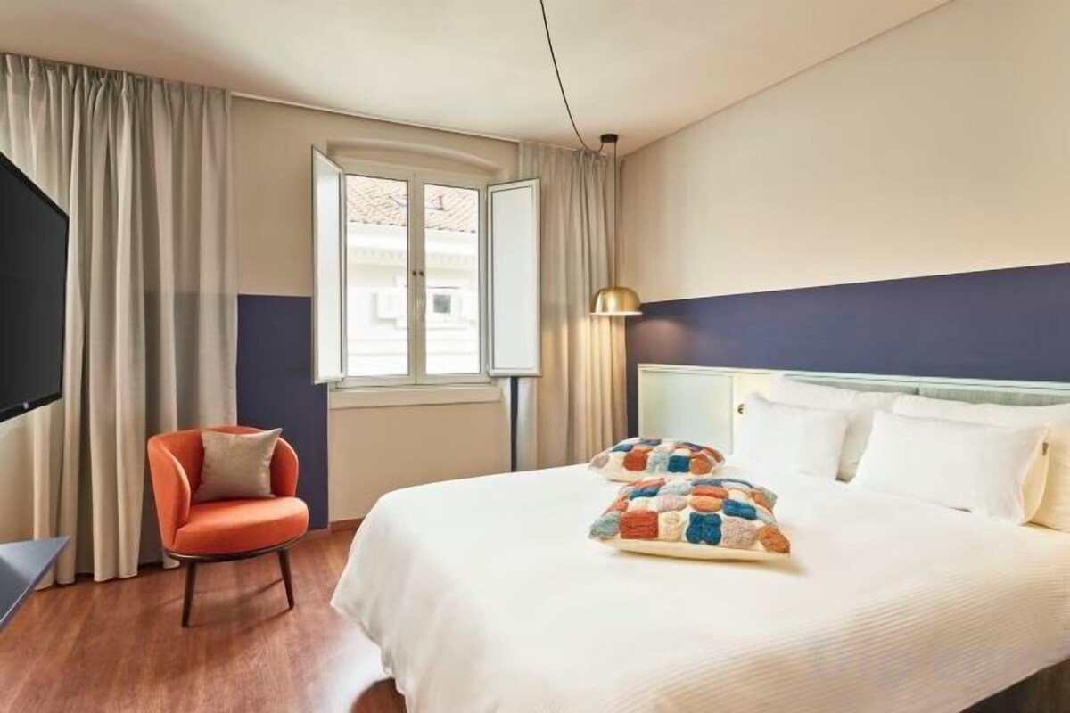 A hotel room with a bed, a chair, a window, and a lamp.