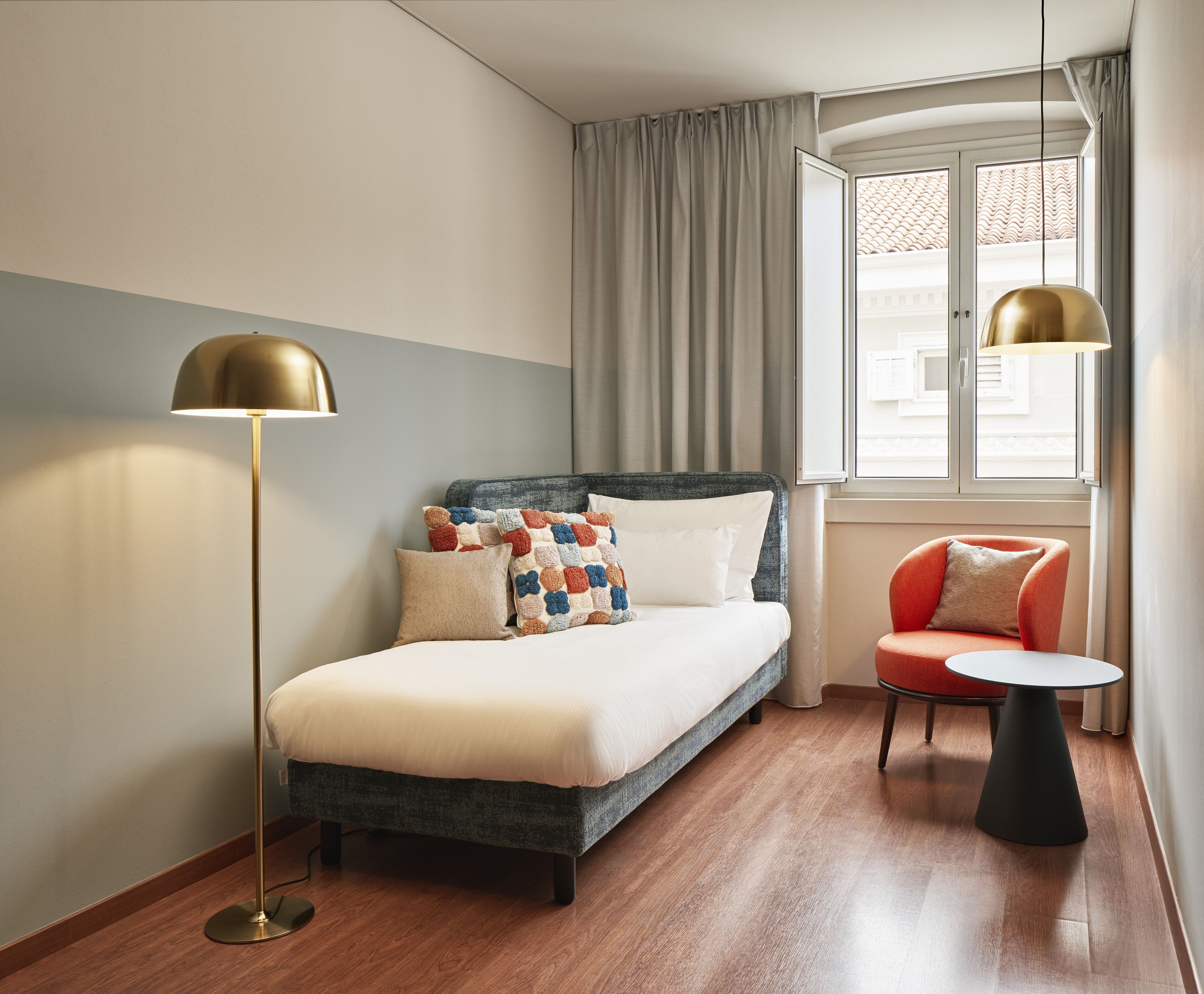 A modern hotel room with a bed, a bedside table, a chair, a floor lamp, and a window with curtains.
