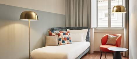 A modern hotel room with a bed, a bedside table, a chair, a floor lamp, and a window with curtains.