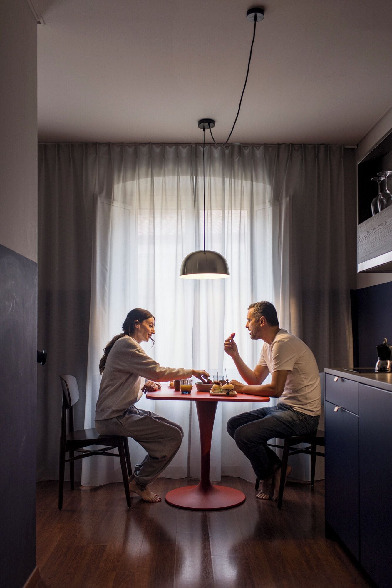 Un coin repas avec une table ronde, deux chaises, un luminaire suspendu et une vue à travers des rideaux légers.