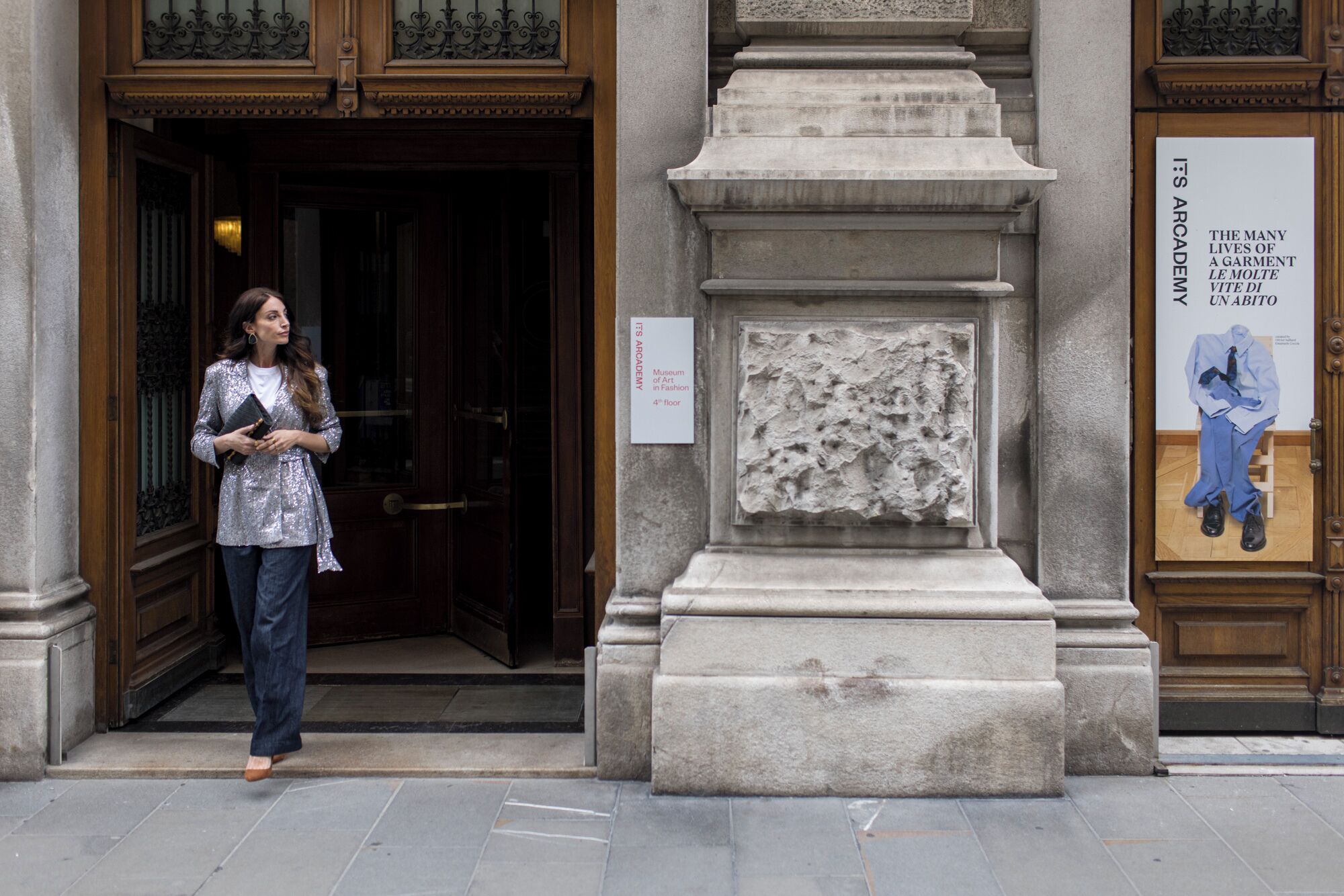 A woman standing in front of a building with a stone facade and a sign that reads "Museum of Art in Fashion 4th floor".