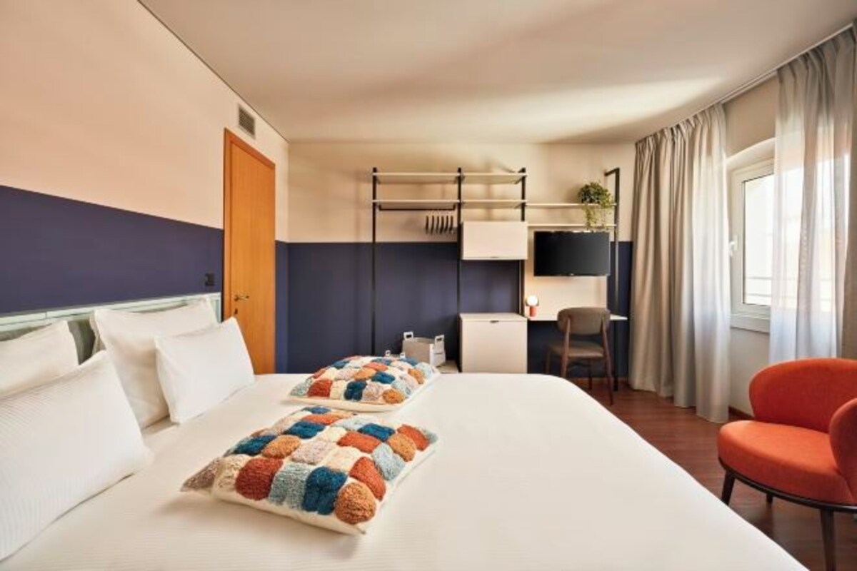 A hotel room with a bed, two pillows, a desk, a chair, a TV, and a window with curtains.