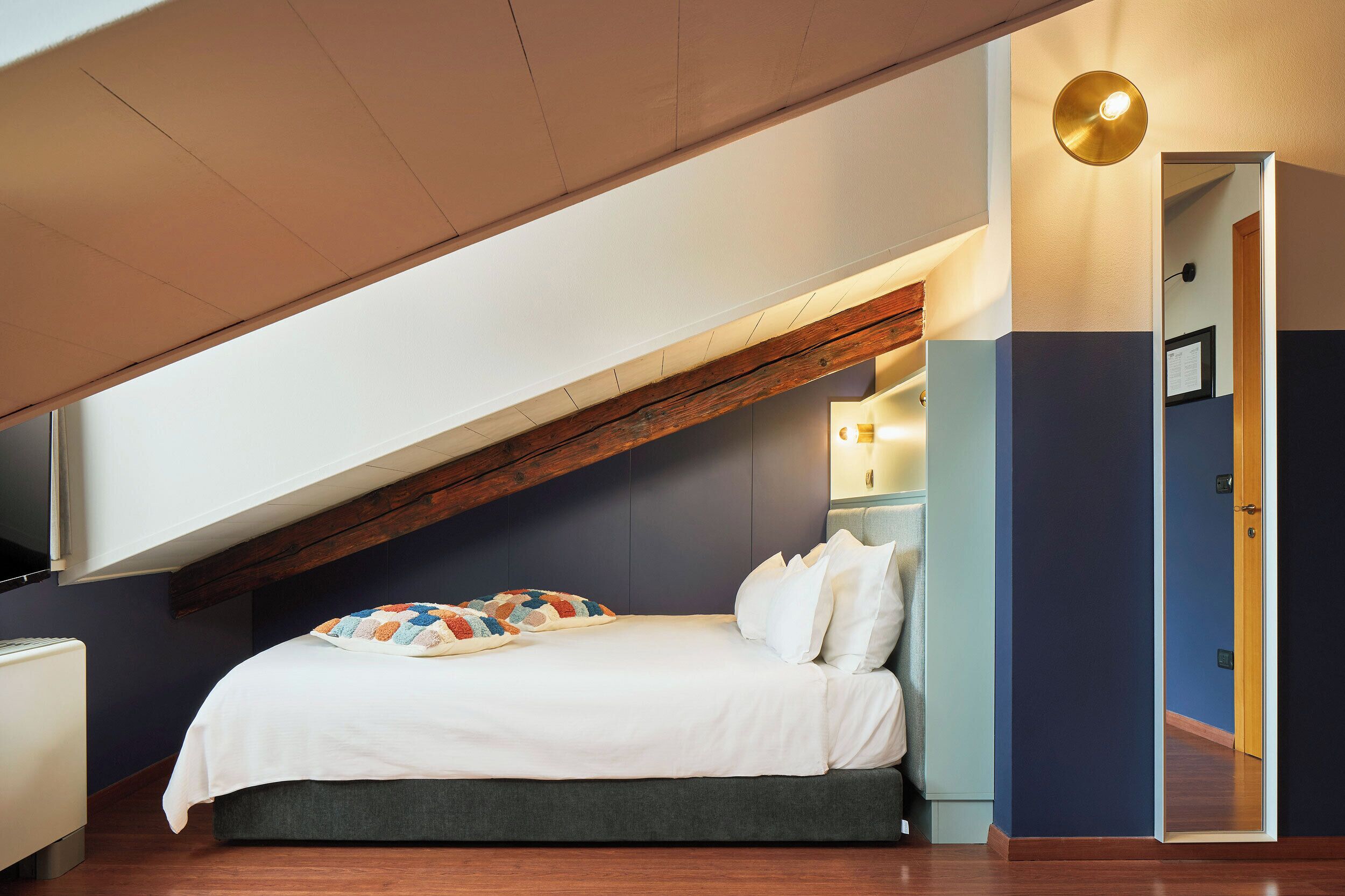 A bedroom with a sloped ceiling, a bed with white bedding, a colorful throw blanket, and a nightstand with a lamp.