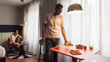 Camera d'albergo moderna con angolo ristoro, un tavolo rosso, cibi per la colazione e un laptop su una sedia.