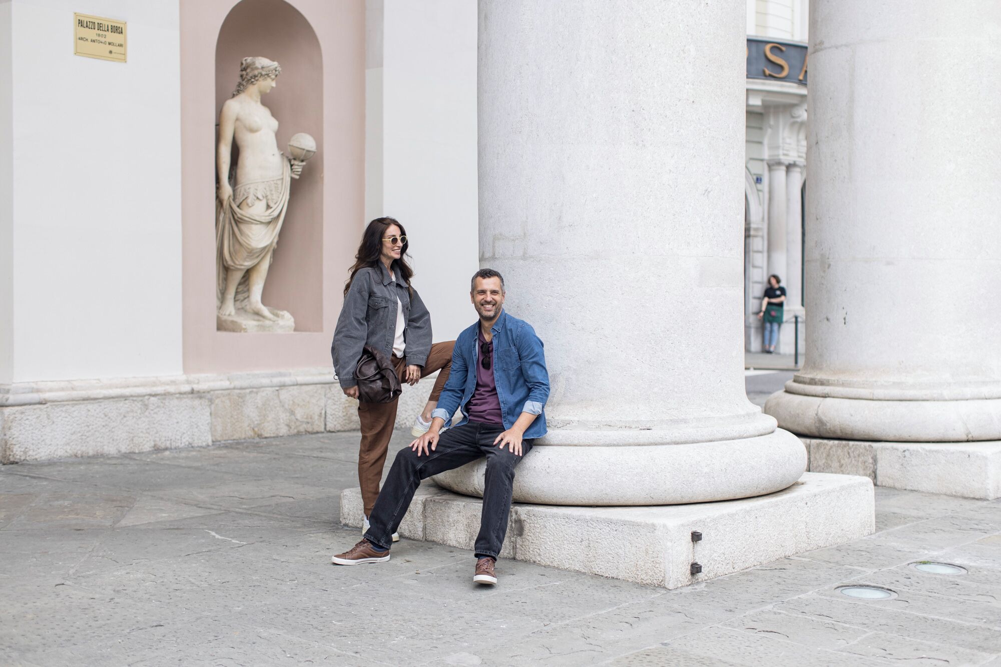 Un uomo e una donna seduti su una panchina di pietra di fronte a un edificio classico con colonne e una statua.