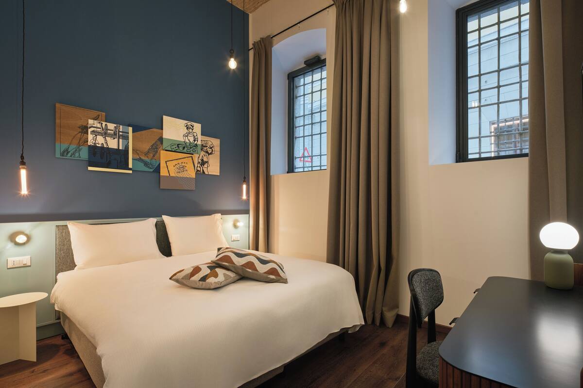 A hotel room with a bed, a desk, a chair, and artwork on the wall.